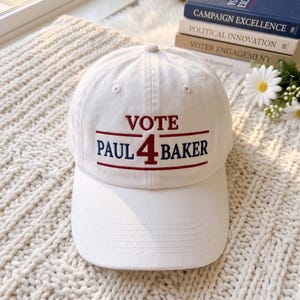 May include: A white baseball cap featuring embroidered text in red and blue. The word VOTE is stitched in red above a horizontal red line. Below this, the text PAUL 4 BAKER is embroidered in blue, with the number 4 in red, followed by another red line.