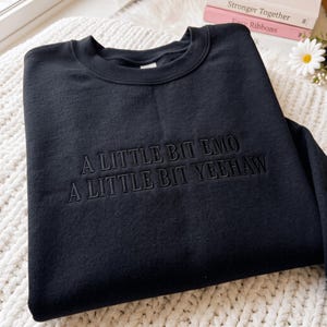 May include: A black crewneck sweatshirt featuring the embroidered text A LITTLE BIT EMO A LITTLE BIT YEEHAW in a matching black thread. The garment has long sleeves and a ribbed collar, showcasing a monochromatic design with bold lettering.