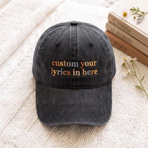 Può includere: Un cappellino da baseball grigio antracite con finitura slavata, caratterizzato dal testo ricamato custom your lyrics in here in fili color arancione spento e azzurro. Il cappellino presenta una corona strutturata e una visiera curva con cuciture.