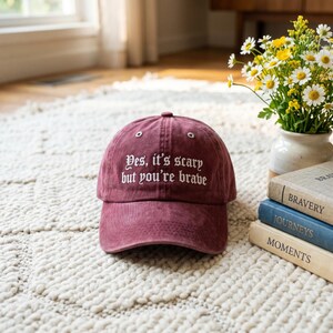 May include: A maroon baseball cap with the embroidered text "Yes, it's scary but you're brave." The hat is sitting on a cream-colored rug. A vase of flowers and a stack of books are in the background.