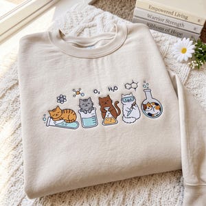 May include: A beige crewneck sweatshirt featuring an embroidered graphic of five cats interacting with laboratory glassware and scientific symbols, including O2 and H2O text. The cats are depicted in various colours and poses with beakers.