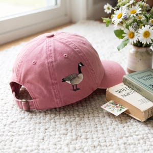 Puede incluir: Una gorra de béisbol de color rosa empolvado con un ganso canadiense bordado en el panel lateral. La gorra cuenta con una corona estructurada, una visera curva y una correa ajustable con hebilla de metal en la parte posterior para un ajuste seguro.