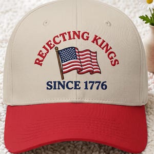 Peut inclure: Casquette de baseball avec visi&egrave;re et bouton rouges, et couronne beige. Le texte brod&eacute; sur la casquette dit "REJECTING KINGS" et "SINCE 1776" avec un drapeau am&eacute;ricain.