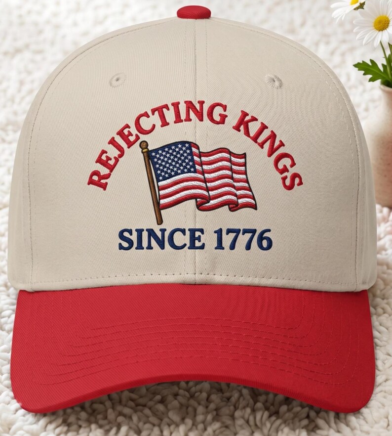 Peut inclure: Casquette de baseball avec visi&egrave;re et bouton rouges, et couronne beige. Le texte brod&eacute; sur la casquette dit "REJECTING KINGS" et "SINCE 1776" avec un drapeau am&eacute;ricain.