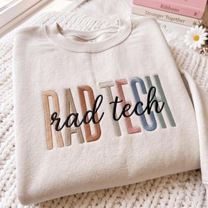 以下が含まれることがあります： クリーム色のクルーネックトレーナーで、RAD TECHという刺繍が施されています。RADという文字は様々な落ち着いた色合いのブロック体で刺繍され、その上にtechという文字が黒い筆記体の刺繍で重ねられています。