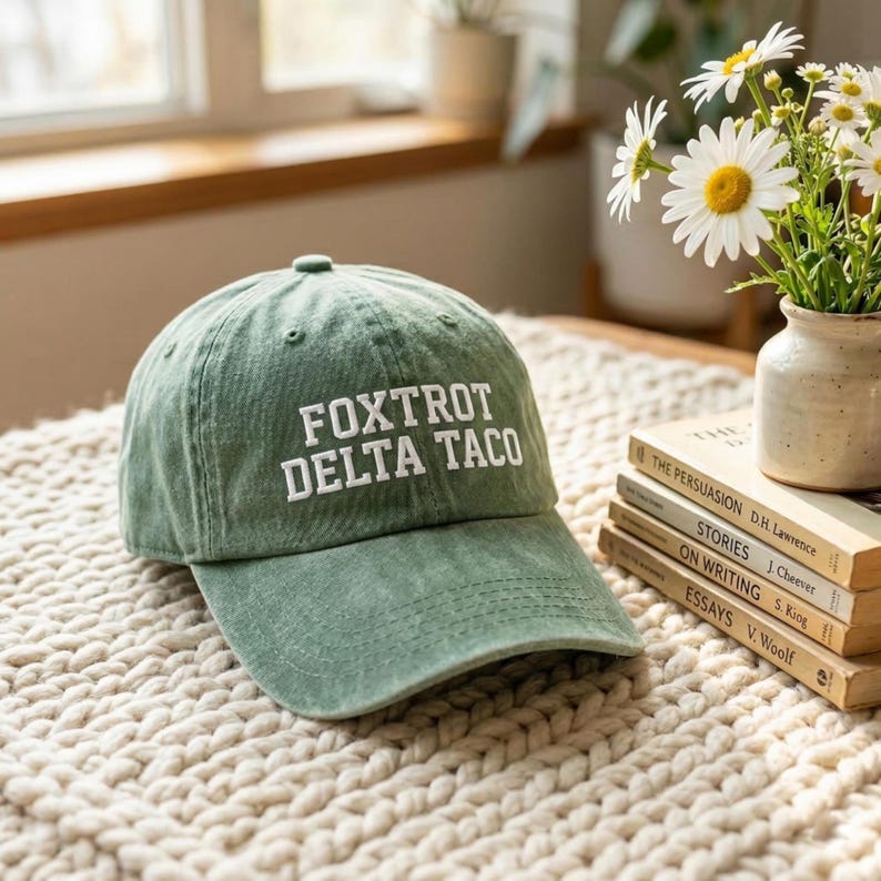 Pu&ograve; includere: Un cappellino da baseball verde con la scritta "FOXTROT DELTA TACO" ricamata in bianco. Il cappello &egrave; appoggiato su una superficie strutturata color crema. Un piccolo vaso di margherite e una pila di libri sono sullo sfondo.