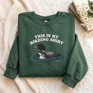 May include: A dark green crewneck sweatshirt featuring a graphic of a loon wearing binoculars on the water. The text "THIS IS MY BIRDING SHIRT" is printed in white above the bird. The sweatshirt has long sleeves and a ribbed collar and cuffs.