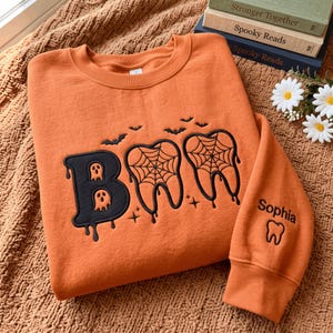 Könnte beinhalten: Ein orangefarbenes Sweatshirt mit Rundhalsausschnitt und schwarzer Stickerei, die das Wort BOO bildet, wobei die beiden O durch zahnförmige Designs mit Spinnennetzen ersetzt sind. Das Motiv enthält kleine Geister und Fledermäuse. Der Ärmel zeigt den Namen Sophia und einen Zahn.