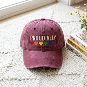 May include: A maroon baseball cap with the text "PROUD ALLY" embroidered in white. Below the text are six embroidered hearts in rainbow colors. The hat is sitting on a white, textured surface.