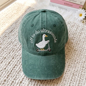 May include: A forest green baseball cap featuring an embroidered white goose riding a grey scooter. The text "off to do ultrasounds" is arched above the goose in white thread. The cap has a structured crown, a curved brim, and stitched eyelets.