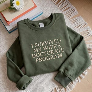 May include: An olive green crewneck sweatshirt featuring the cream-colored text "I SURVIVED MY WIFE'S DOCTORATE PROGRAM" printed in a bold, sans-serif font across the chest. The garment has long sleeves with ribbed cuffs and a ribbed neckline.
