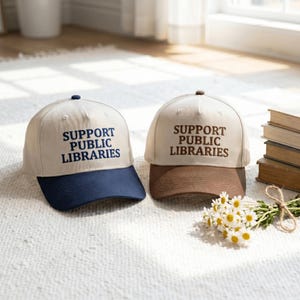May include: Two baseball caps with beige crowns and embroidered text reading SUPPORT PUBLIC LIBRARIES. One cap features a navy blue brim, while the other has a brown brim. Both caps have a button on top and structured panels.