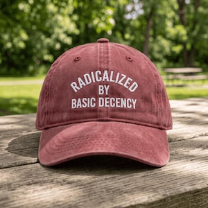 Può includere: Un berretto da baseball rosso mattone sbiadito con visiera curva e un bottone sulla parte superiore. La parte anteriore presenta un testo ricamato in bianco che recita RADICALIZED BY BASIC DECENCY in un arco sopra le parole BASIC DECENCY.