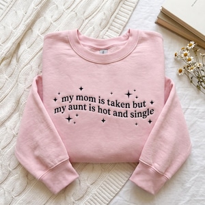 May include: A light pink crewneck sweatshirt featuring dark purple embroidered text that reads, "my mom is taken but my aunt is hot and single," surrounded by small embroidered star icons. The garment has long sleeves and ribbed cuffs.