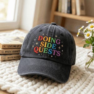 May include: A charcoal gray baseball cap featuring the embroidered text DOING SIDE QUESTS in a rainbow gradient font, surrounded by small star icons. The cap has a structured crown, a curved brim, and a button on top, with a distressed finish.