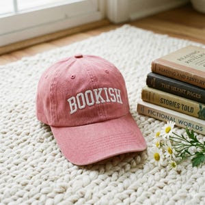 May include: A dusty rose baseball cap with the word "BOOKISH" embroidered in white block letters. The cap is sitting on a white, textured rug next to a stack of books with titles like "Reading and Revelry" and "Fictional Worlds".
