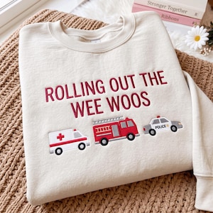 Puede incluir: Una sudadera de cuello redondo color crema con el texto bordado en rojo "ROLLING OUT THE WEE WOOS" sobre tres vehículos de emergencia cosidos: una ambulancia blanca con una cruz roja, un camión de bomberos rojo y un coche de policía.