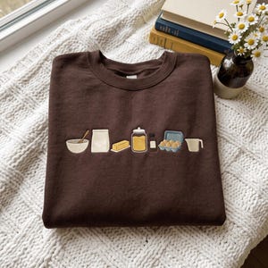 May include: A dark brown crewneck sweatshirt featuring an embroidered horizontal row of baking ingredients including a mixing bowl with a spoon, a bag of flour, a stick of butter, a glass jar, a small bottle, a carton of eggs, and a measuring cup.