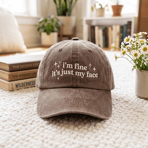 May include: A brown baseball cap with a distressed finish featuring white embroidered text that reads "i'm fine it's just my face" surrounded by small star icons. The cap has a curved brim and a structured front panel with ventilation eyelets.