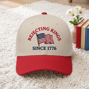Peut inclure: Casquette de baseball avec une visi&egrave;re et un bouton rouges, et une couronne cr&egrave;me. Le texte "REJECTING KINGS SINCE 1776" et un drapeau am&eacute;ricain sont brod&eacute;s. La casquette est pos&eacute;e sur un tapis blanc.