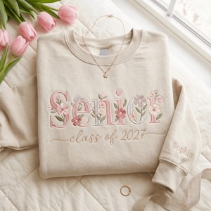 May include: A beige crewneck sweatshirt featuring floral embroidery that spells out the word Senior in pink and light blue tones. Below the word, the text class of 2027 is embroidered in a cursive font. The name Sophia is embroidered on the sleeve.