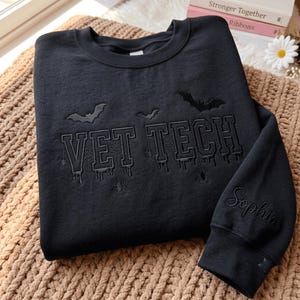 May include: A black crewneck sweatshirt featuring black embroidery that reads VET TECH in a collegiate font with dripping effects. The design includes embroidered bats, spiderwebs, and spiders. The name Sophia is embroidered on the sleeve cuff.