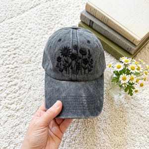 Puede incluir: Una gorra de béisbol gris carbón con un acabado desgastado y lavado. El panel frontal presenta diseños florales bordados en negro, incluyendo girasoles y flores silvestres. La gorra tiene una visera curva con costuras visibles.