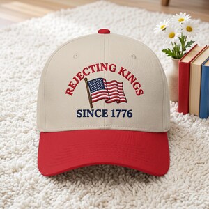 Peut inclure: Casquette de baseball avec une visi&egrave;re et un bouton rouges, et une couronne cr&egrave;me. La casquette porte l'inscription "REJECTING KINGS SINCE 1776" avec un drapeau am&eacute;ricain brod&eacute;. La casquette est pos&eacute;e sur un tapis blanc.