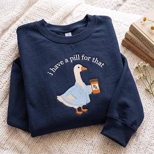 May include: A navy blue crewneck sweatshirt featuring an embroidered graphic of a white goose wearing a light blue medical coat and holding an orange prescription pill bottle. The text "i have a pill for that" is embroidered in white above the goose.