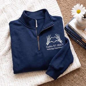 May include: A navy blue quarter-zip sweatshirt featuring a white graphic of two skeletal hands forming a heart shape. Below the graphic, the text reads "Sophia R.T. (R)(CT) Radiology Department" in white lettering. The garment has a metal zipper.