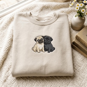 May include: A beige crewneck sweatshirt featuring an embroidered graphic of two pug dogs sitting side by side. One pug is fawn-colored with a black muzzle, and the other pug is solid black. The design is centered on the chest of the garment.