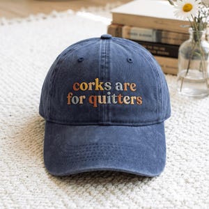 Puede incluir: Una gorra de béisbol azul marino con un acabado de tela desgastada. La parte frontal de la gorra presenta un texto bordado en naranja y azul claro que dice "corks are for quitters", con una corona estructurada y una visera curva.