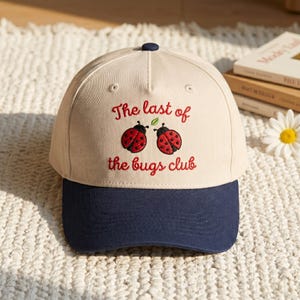 May include: A baseball cap with a cream-colored crown and a navy blue brim. The cap features the embroidered text "The last of the bugs club" with two red ladybug designs. A navy blue button is on top.