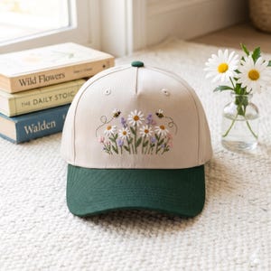 May include: A beige baseball cap with a dark green brim, featuring an embroidered design of white daisies, purple lavender, and two small bees on the front panel. The cap has a structured crown and a button at the top centre of the fabric.
