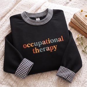 May include: A black sweatshirt featuring a black and white gingham collar and cuffs. The text "occupational therapy" is embroidered across the chest in a multi-colored font with orange, yellow, and blue letters.