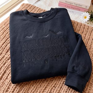 May include: A black crewneck sweatshirt featuring tonal black embroidery that reads "WELCOME TO NIGHT SHIFT" in a collegiate font, accented with embroidered bats and a dripping effect. The sleeve is personalized with the name "Sophia" in script.