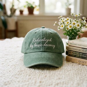 Peut inclure: Casquette de baseball vert sauge avec la phrase "Radicalized by basic decency" brodée en blanc. Le chapeau est présenté sur une surface texturée, avec des livres et des fleurs en arrière-plan.