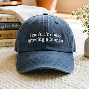 May include: A blue baseball cap with a distressed finish featuring white embroidered text on the front that reads, i can't. i'm busy growing a human. The cap has a structured crown, a curved brim, and a button at the top centre of the fabric panels.