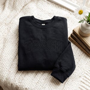 Könnte beinhalten: Ein schwarzes Sweatshirt mit Rundhalsausschnitt, auf dessen Brust das Wort TEACH in passendem schwarzem Garn in einer College-Schriftart gestickt ist. Der Ärmel ist mit dem Namen Sophia in Schreibschrift ebenfalls in Schwarz bestickt.