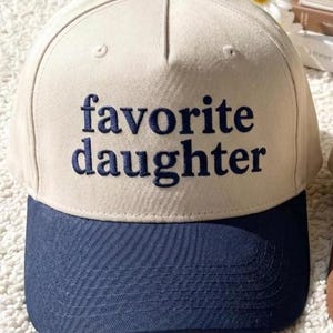 Puede incluir: Gorra de b&eacute;isbol con corona color crema y visera azul marino. Las palabras "favorite daughter" est&aacute;n bordadas en azul marino en la parte delantera. La gorra tiene un bot&oacute;n azul marino en la parte superior.