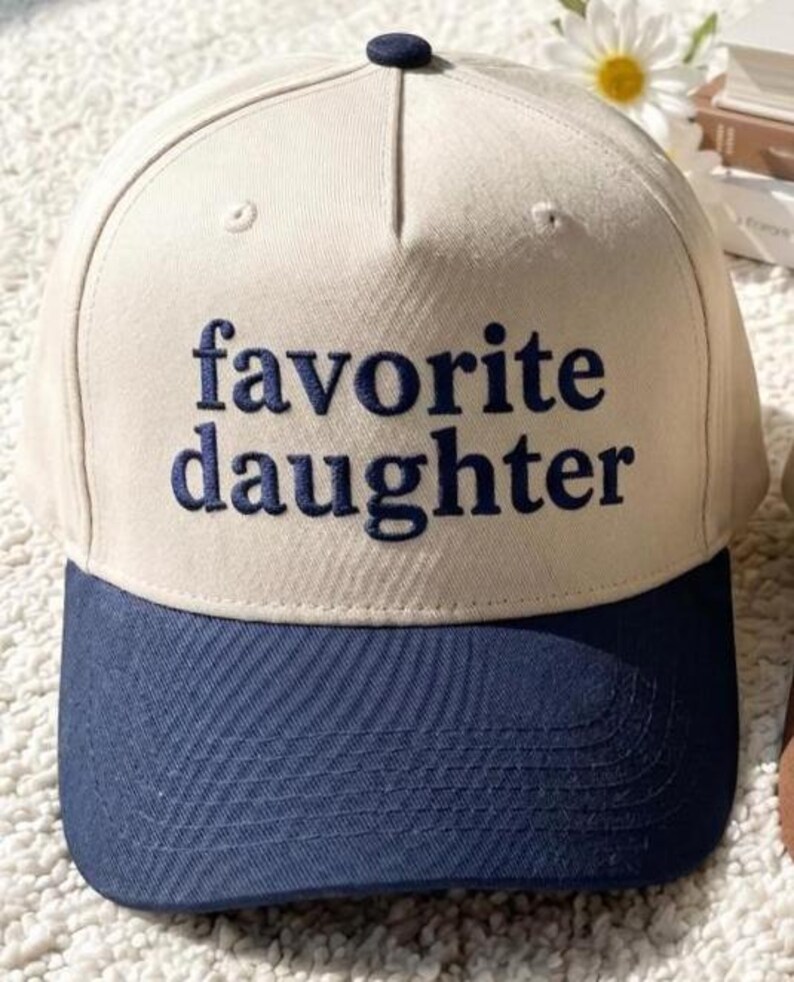 Puede incluir: Gorra de b&eacute;isbol con corona color crema y visera azul marino. Las palabras "favorite daughter" est&aacute;n bordadas en azul marino en la parte delantera. La gorra tiene un bot&oacute;n azul marino en la parte superior.