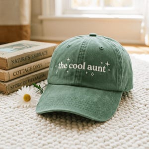 May include: A sage green baseball cap featuring the white embroidered text "the cool aunt" centered on the front panel, surrounded by small white star-shaped accents. The cap has a structured crown, a curved brim, and visible stitching details.