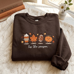 以下が含まれることがあります： コーヒーカップ、カエデの葉、アメリカンフットボール、カボチャのキャラクターが手をつないでいる刺繍が施されたダークブラウンのクルーネックトレーナー。キャラクターの下には白い糸で「tis the season」という文字と小さな葉が刺繍されています。