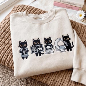 May include: A cream-colored crewneck sweatshirt featuring an embroidered graphic of four black cats performing medical tasks. The cats are shown holding an X-ray of a hand, a skeletal X-ray, an ultrasound machine, and operating a CT scanner.