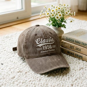 May include: A brown baseball cap with white embroidered text on the front that reads Classic 1956 Limited Edition. The cap features a curved brim, a button on the top, and stitched eyelets for ventilation, showcasing a weathered fabric texture.