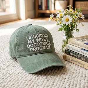 May include: A sage green baseball cap featuring the white embroidered text I SURVIVED MY WIFE'S DOCTORATE PROGRAM on the front panel. The cap has a structured crown, a curved brim, and stitched eyelets for ventilation, presenting a casual style.