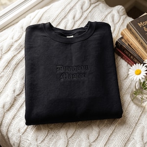 Puede incluir: Una sudadera negra de cuello redondo con las palabras Dungeon Master bordadas en hilo negro a juego en el centro del pecho. La prenda presenta un diseño clásico de manga larga con cuello, puños y dobladillo acanalados en color negro.