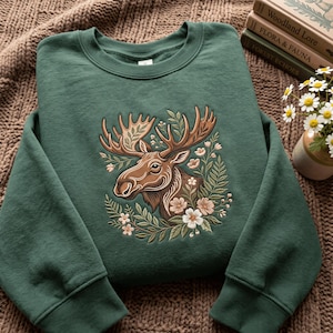 Könnte beinhalten: Ein waldgrüner Rundhals-Pullover mit einer Stickerei eines braunen Elchkopfes, der von komplizierten Blumen- und Farnmustern in Creme-, Hellrosa- und Grüntönen umgeben ist und mittig auf der Vorderseite des Kleidungsstücks platziert ist.