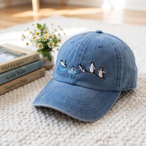 Può includere: Un berretto da baseball in denim blu con un ricamo raffigurante cinque pinguini in varie pose, tra cui uno che si tuffa nell'acqua blu, uno che sguazza e tre in piedi su una sottile linea nera sui pannelli frontali del cappello.