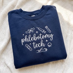Puede incluir: Una sudadera azul marino de cuello redondo con el texto bordado en blanco phlebotomy tech. El diseño incluye iconos de línea blanca de una jeringa, un tubo de ensayo, una tirita, un torniquete, una bola de algodón y un brazo con aguja.